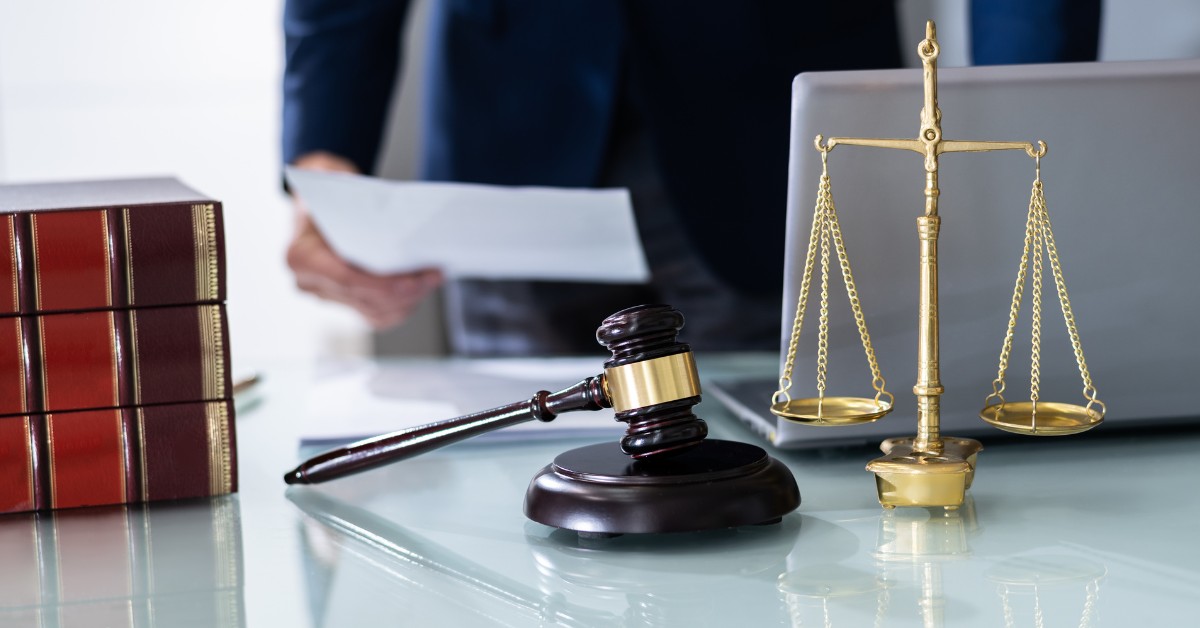A Person Standing Behind A Desk With A Piece of Paper In Their Hand With A Laptop, Law Books, Gavel Hammer, and Scales of Justice on the Table | Does A Revocable Trust Protect You From Liability | The TGQ Law Firm